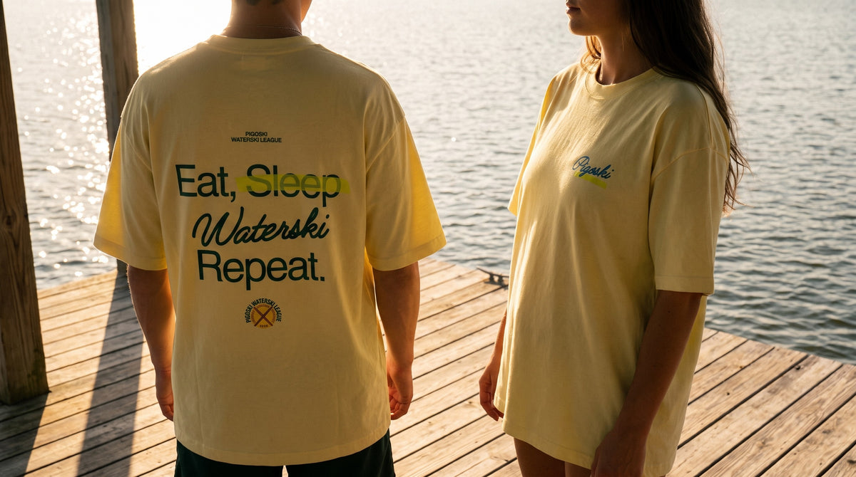 Two models on a boathouse dock at golden hour wearing the PIGOSKI Ritual Tee — Tees collection banner