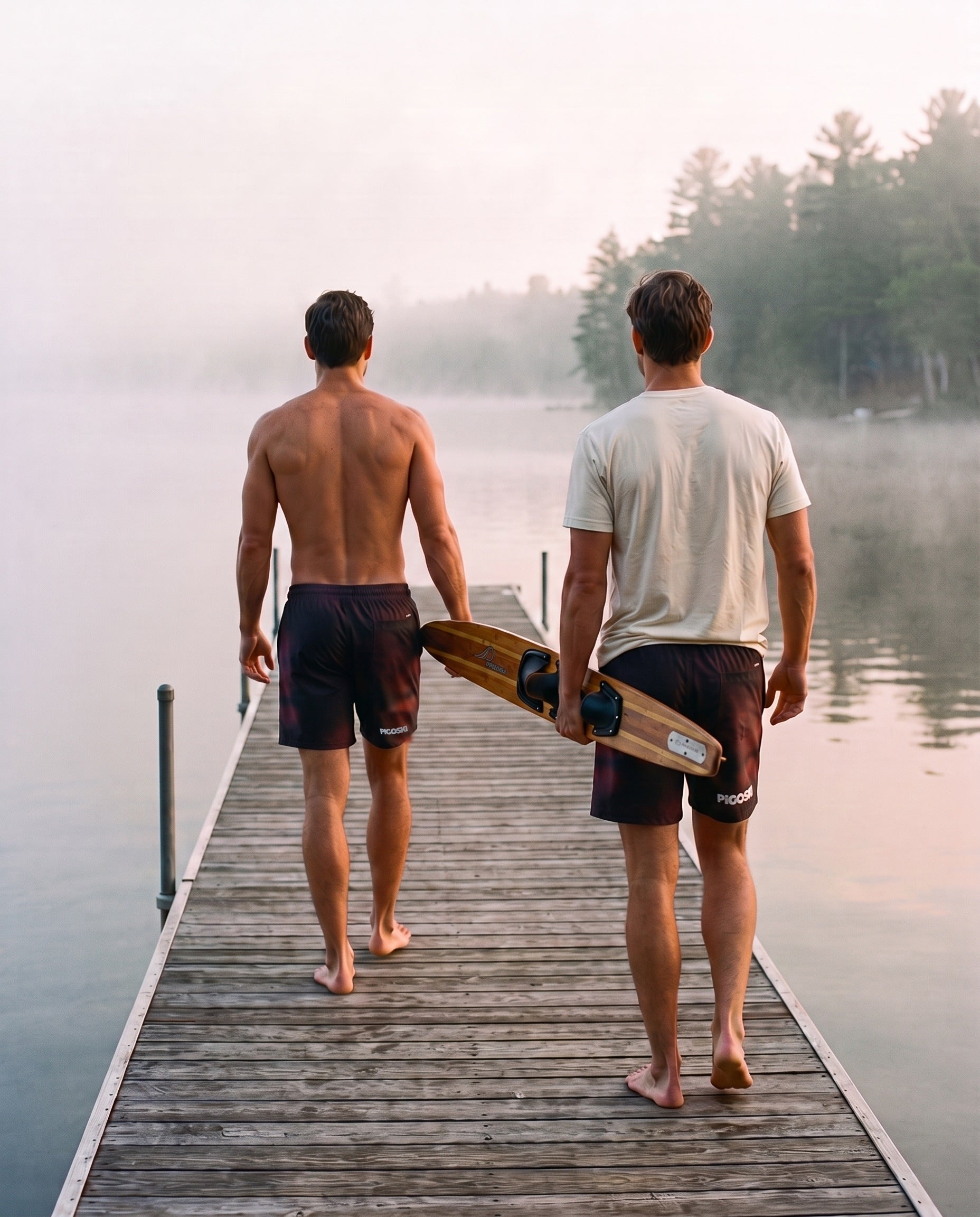 Two skiers in PIGOSKI board shorts carrying a wooden slalom ski down the dock